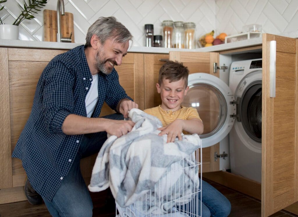An adult and a child are loading a blanket into a washing machine in a kitchen laundry area, both smiling.