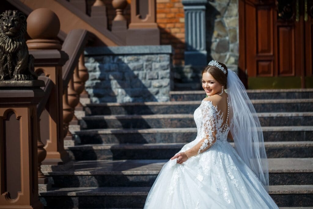 A bride in a white lace wedding dress and veil stands on outdoor stone steps, smiling and holding her skirt.